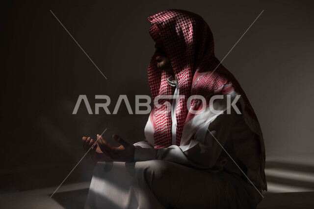 التوبة والرجوع الى الله، الاجتهاد في العباده والطاعة في شهر رمضان المبارك, مفهوم الدعاء والترجي والطلب، بورتريه لشاب عربي خليجي سعودي يرتدي الشماغ والثوب التقليدي يجلس على الأرض يرفع يديه ويدعو الله بإيماءات تدل على التوسل والتضرع، خلفية سوداء