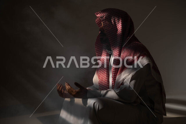 مفهوم الدعاء والترجي والطلب، بورتريه لشاب عربي خليجي سعودي يرتدي الشماغ والثوب التقليدي يجلس على الأرض يرفع يديه ويدعو الله بإيماءات تدل على التوسل والتضرع، التوبة والرجوع الى الله، الاجتهاد في العباده والطاعة في شهر رمضان المبارك, خلفية سوداء
