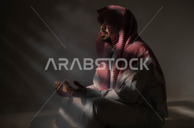 الاجتهاد في العباده والطاعة في شهر رمضان المبارك, مفهوم الدعاء والترجي والطلب، بورتريه لشاب عربي خليجي سعودي يرتدي الشماغ والثوب التقليدي يجلس على الأرض يرفع يديه ويدعو الله بإيماءات تدل على التوسل والتضرع، التوبة والرجوع الى الله، خلفية سوداء