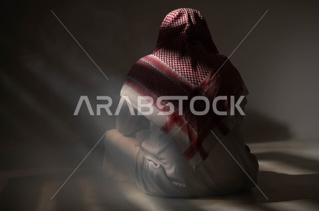الإنعزال النفسي والشخصية الانطوائية، معاقبة النفس وتأنيب الضمير، الآثار السلبية للادمان والتعاطي، المعاناة من الاضطرابات النفسية، بورتريه من الخلف لرجل عربي خليجي سعودي يرتدي الزي التقليدي يجلس على الارض مصاب بالإكتئاب والإنهيار العصبي، خلفية سوداء