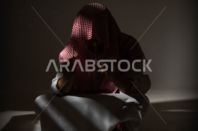 المعاناة من الاضطرابات النفسية والمشاكل الصحية، الادمان على التبغ والمخدرات، بورتريه لرجل عربي خليجي سعودي يرتدي الشماغ والثوب التقليدي يجلس على الأرض يمسك بيديه عقب سيجارة، عادات سلوكية ضارة وتعاطي مواد سامة، معاقبة النفس وتأنيب الضمير، خلفية سوداء