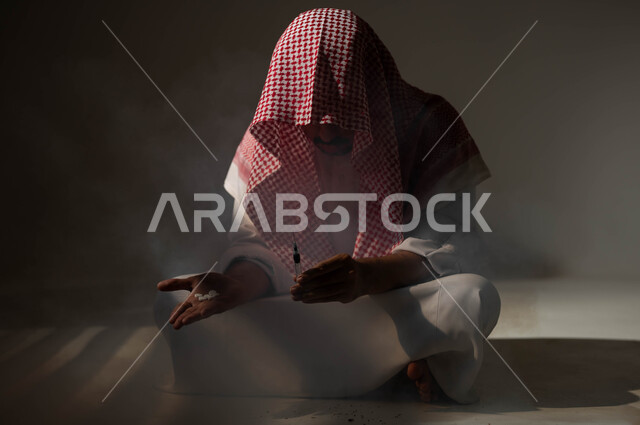 الأثار السلبية للادمان على التبغ والمخدرات، عادات سلوكية ضارة وتعاطي مواد سامة، المعاناة من الاضطرابات النفسية والمشاكل الصحية، بورتريه لرجل عربي خليجي سعودي يرتدي الشماغ والثوب التقليدي يجلس على الأرض يمسك بيديه الحقن والاقراص المخدرة، خلفية سوداء