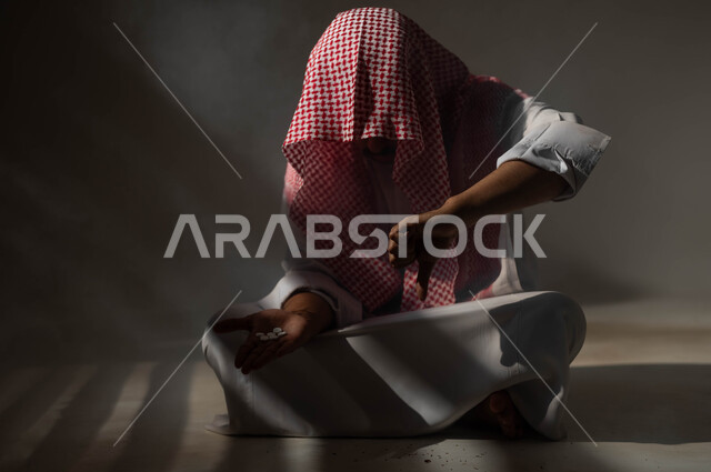 ايماءات وتعابير الرفض وعدم الرضا، عادات سلوكية ضارة وتعاطي مواد سامة، الادمان على التبغ والمخدرات، المعاناة من الاضطرابات النفسية والمشاكل الصحية، بورتريه لرجل عربي خليجي سعودي يرتدي الزي التقليدي يجلس على الأرض يمسك بيده الاقراص المخدرة، خلفية سوداء
