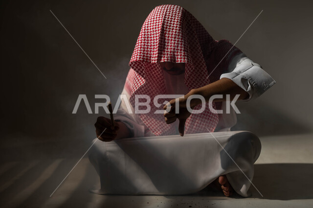 Gestures and expressions of rejection and dissatisfaction, harmful behavioral habits and the use of harmful and toxic substances, addiction to tobacco and drugs, suffering from psychological disorders and health problems, a portrait of a Saudi Gulf Arab man wearing traditional clothing, sitting on the floor holding a cigarette butt in his hand, black background.