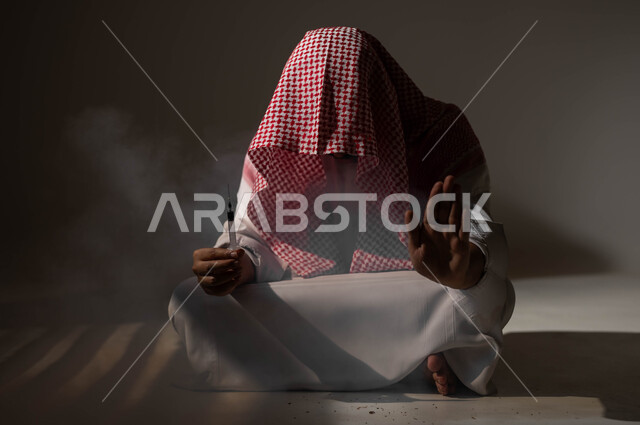 عادات سلوكية ضارة وتعاطي مواد سامة، طلب التوقف والامتناع عن الادمان على التبغ والمخدرات، المعاناة من الاضطرابات النفسية والمشاكل الصحية، بورتريه لرجل عربي خليجي سعودي يرتدي الزي التقليدي يجلس على الأرض يمسك بيده جرعة وحقنة مخدرة، خلفية سوداء