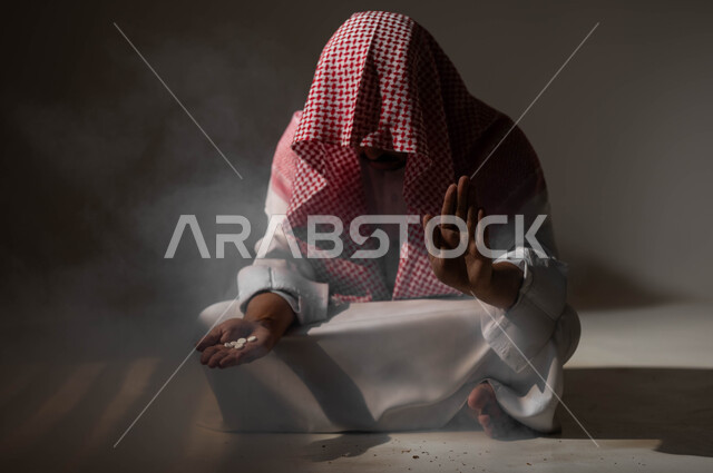 طلب التوقف والامتناع عن الادمان على التبغ والمخدرات، المعاناة من الاضطرابات النفسية والمشاكل الصحية، بورتريه لرجل عربي خليجي سعودي يرتدي الزي التقليدي يجلس على الأرض يمسك بيده الاقراص المخدرة، عادات سلوكية ضارة وتعاطي مواد سامة، خلفية سوداء