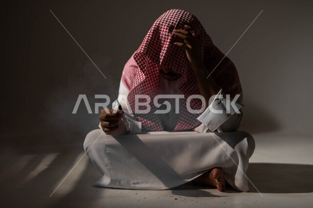 المعاناة من الاضطرابات النفسية والمشاكل الصحية، بورتريه لرجل عربي خليجي سعودي يرتدي الشماغ والثوب التقليدي يجلس على الأرض يمسك بيده عقب سيجارة، الادمان على التبغ والمخدرات، عادات سلوكية ضارة وتعاطي مواد سامة، معاقبة النفس وتأنيب الضمير، خلفية سوداء