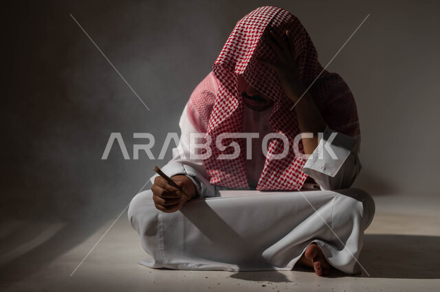 المعاناة من الاضطرابات النفسية والمشاكل الصحية، بورتريه لرجل عربي خليجي سعودي يرتدي الشماغ والثوب التقليدي يجلس على الأرض يمسك بيده عقب سيجارة، الادمان على التبغ والمخدرات، عادات سلوكية ضارة وتعاطي مواد سامة، معاقبة النفس وتأنيب الضمير، خلفية سوداء