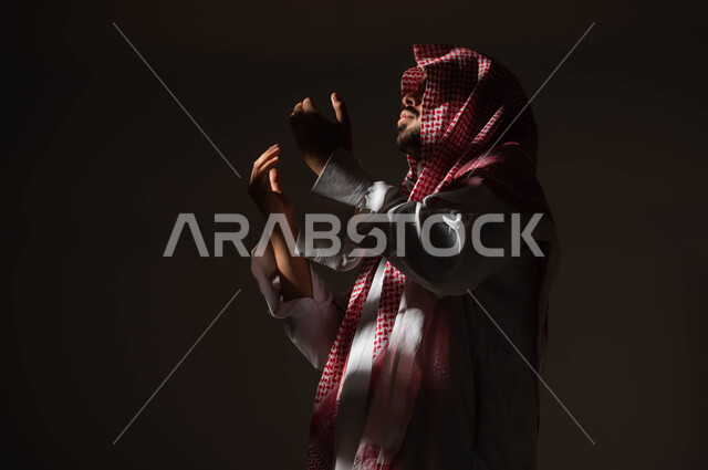 مفهوم الدعاء والترجي والطلب، الاجتهاد في العباده والتقرب الى الله في شهر رمضان المبارك, بورتريه مقرب من الجانب لرجل عربي خليجي سعودي يرتدي الشماغ والثوب التقليدي يرفع يديه ويدعو الله بإيماءات تدل على التوسل والتضرع،  خلفية سوداء