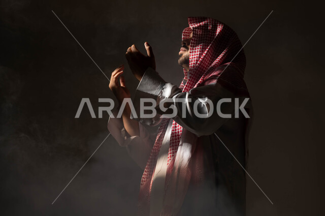 إيماءات تدل على التوسل والتضرع، مفهوم الدعاء والترجي والطلب، الاجتهاد في العباده والتقرب الى الله في شهر رمضان المبارك, بورتريه مقرب من الجانب لرجل عربي خليجي سعودي يرتدي الشماغ والثوب التقليدي يرفع يديه ويدعو الله، خلفية سوداء