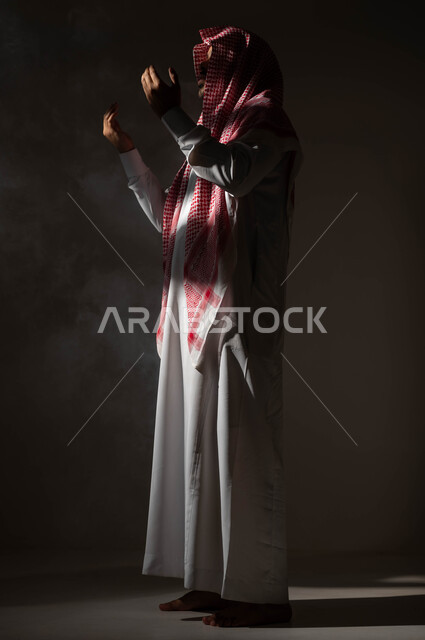 الاجتهاد في العباده والتقرب الى الله في شهر رمضان المبارك, مفهوم الدعاء والترجي والطلب، بورتريه من الجانب لرجل عربي خليجي سعودي يرتدي الشماغ والثوب التقليدي يرفع يديه ويدعو الله بإيماءات تدل على التوسل والتضرع، صورة الجسم بالكامل، خلفية سوداء