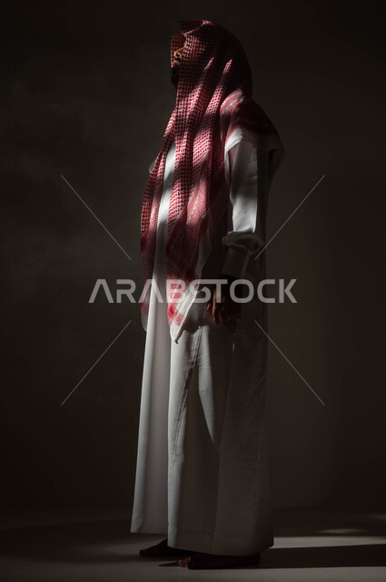 Finding a solution to a problem, feeling bored and tired from the pressures of life and work, contemplating and planning for the future, a side portrait of a Saudi Gulf Arab man wearing the traditional thobe and shemagh, looking at something with gestures of thought, contemplation, concentration and absent-mindedness, full body portrait, black background