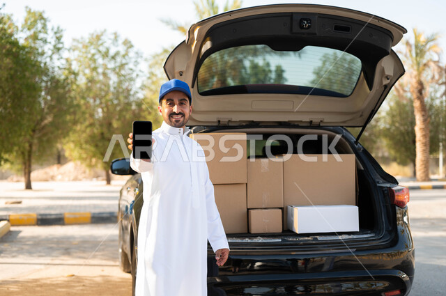 الشراء والتسوق عبر الانترنت، ايماءات الرضا والسرور بتوصيل الطلبات للزبائن، طلب المنتجات عبر تطبيقات الهاتف المحمول، عامل توصيل عربي خليجي سعودي مبتسم يرتدي الثوب التقليدي وقبعة زرقاء يقف بجانب سيارة البضاعة، عرض شاشة سوداء فارغة عبر الهاتف المحمول