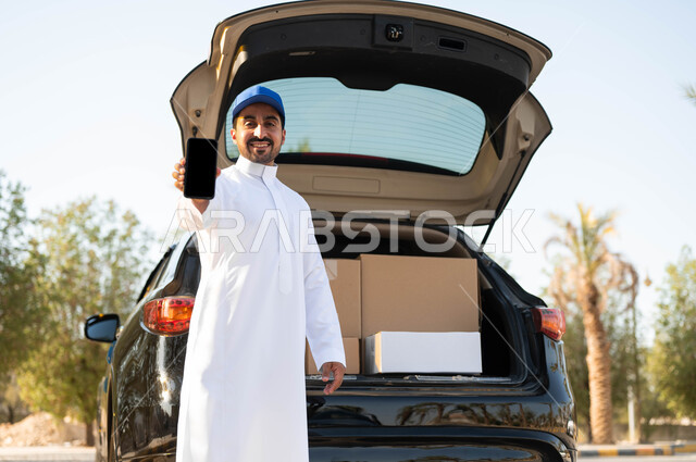 Gestures of satisfaction and pleasure in delivering orders to customers, ordering products via mobile phone applications, a smiling Saudi Arabian Gulf delivery worker wearing a traditional dress and a blue hat standing next to the goods car, shopping and purchasing online, displaying a blank black screen on the mobile phone