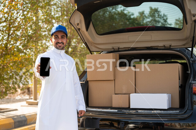 التسوق والشراء عبر الانترنت، عرض شاشة سوداء فارغة عبر الهاتف المحمول، طلب المنتجات عبر تطبيقات الهاتف المحمول، عامل توصيل عربي خليجي سعودي مبتسم يرتدي الثوب التقليدي وقبعة زرقاء يقف بجانب سيارة البضاعة، ايماءات الرضا والسرور بتوصيل الطلبات للزبائن