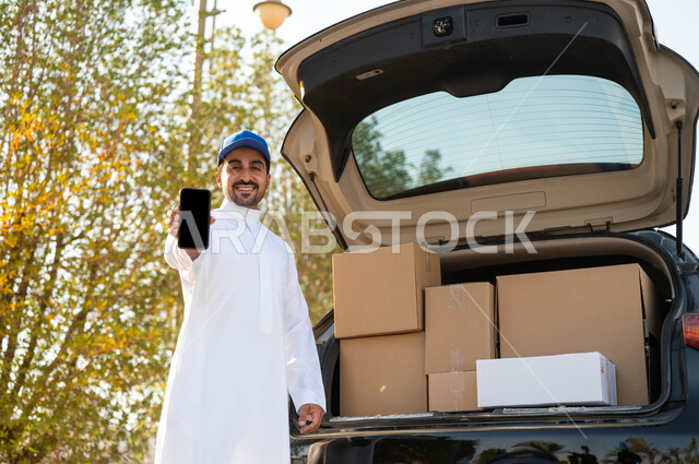 عرض شاشة سوداء فارغة عبر الهاتف المحمول، طلب المنتجات عبر تطبيقات الهاتف المحمول، عامل توصيل عربي خليجي سعودي مبتسم يرتدي الثوب التقليدي وقبعة زرقاء يقف بجانب سيارة البضاعة، التسوق والشراء عبر الانترنت، ايماءات الرضا والسرور بتوصيل الطلبات للزبائن