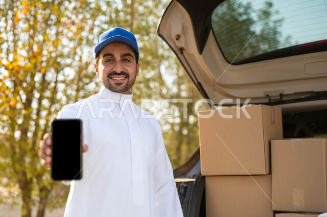 Ordering products via mobile phone applications, a smiling Saudi Arabian Gulf delivery worker wearing a traditional dress and a blue hat standing next to the goods car, displaying a blank black screen via the mobile phone, shopping and purchasing online, gestures of satisfaction and pleasure in delivering orders to customers.