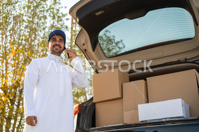 إجراء المكالمات الهاتفية لتأكيد الحجوزات، التسوق الذكي وطلب المنتجات أونلاين، مندوب توصيل عربي خليجي سعودي يرتدي الثوب التقليدي وقبعة زرقاء يقف بجانب سيارة البضائع، التواصل الفعال مع العملاء، توصيل المنتجات السريع