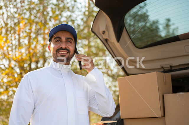 التسوق الذكي وطلب المنتجات أونلاين، مندوب توصيل عربي خليجي سعودي يرتدي الثوب التقليدي وقبعة زرقاء يقف بجانب سيارة البضائع، إجراء المكالمات الهاتفية لتأكيد الحجوزات، التواصل الفعال مع العملاء، توصيل المنتجات السريع