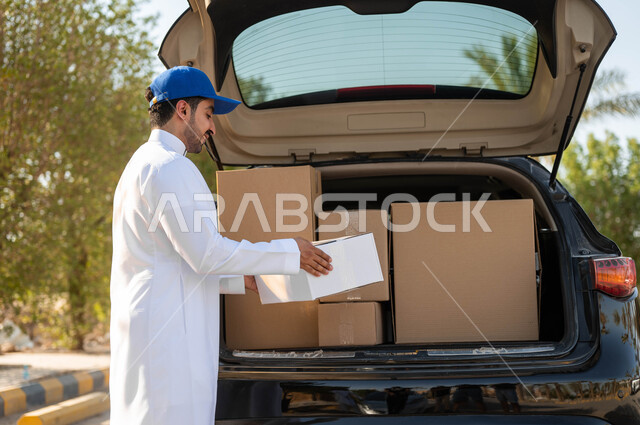 ايماءات الرضا والسرور خلال العمل، طلب المنتجات والتسوق عبر الإنترنت، خدمات الشحن والتوصيل السريع والموثوق، مندوب توصيل عربي خليجي سعودي يرتدي الزي التقليدي وقبعة زرقاء يقوم بأخذ صندوق كرتون أبيض من السيارة، عملية تسليم الطرود للمنازل
