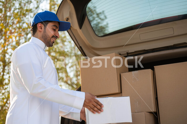 خدمات الشحن والتوصيل السريع والموثوق، مندوب توصيل عربي خليجي سعودي يرتدي الزي التقليدي وقبعة زرقاء يقوم بأخذ صندوق كرتون أبيض من السيارة، طلب المنتجات والتسوق عبر الإنترنت، عملية تسليم الطرود للمنازل، ايماءات الرضا والسرور خلال العمل
