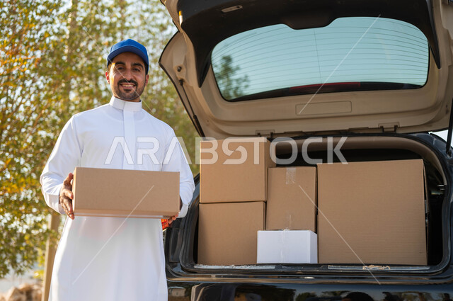 التسوق وطلب المنتجات عبر الإنترنت، عملية توصيل الطلبات للزبائن، شاب عربي خليجي سعودي يرتدي الزي التقليدي وقبعة زرقاء يعمل كمندوب مبيعات ويحمل بيده صندوق كرتوني، النظر الى الكاميرا بإيماءات السعادة والسرور، الشحن والتوصيل ذات الجودة العالية