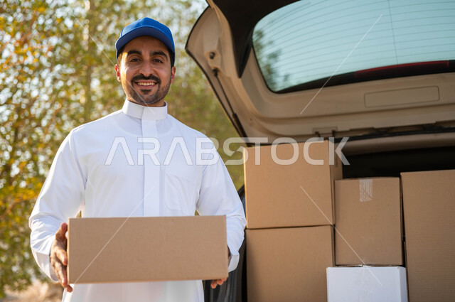 النظر الى الكاميرا بإيماءات السعادة والسرور، الشحن والتوصيل ذات الجودة العالية، عملية توصيل الطلبات للزبائن، شاب عربي خليجي سعودي يرتدي الزي التقليدي وقبعة زرقاء يعمل كمندوب مبيعات ويحمل بيده صندوق كرتوني، التسوق وطلب المنتجات عبر الإنترنت