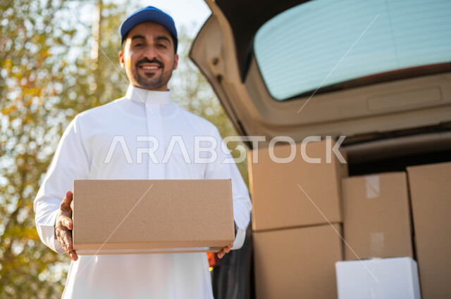 الشحن والتوصيل ذات الجودة العالية، عملية توصيل الطلبات للزبائن، شاب عربي خليجي سعودي يرتدي الزي التقليدي وقبعة زرقاء يعمل كمندوب مبيعات ويحمل بيده صندوق كرتوني، النظر الى الكاميرا بإيماءات السعادة والسرور، التسوق وطلب المنتجات عبر الإنترنت