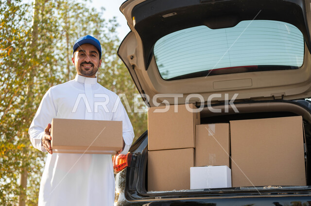 عملية توصيل الطلبات للزبائن، الشحن والتوصيل ذات الجودة العالية، شاب عربي خليجي سعودي يرتدي الزي التقليدي وقبعة زرقاء يعمل كمندوب مبيعات ويحمل بيده صندوق كرتوني، النظر الى الكاميرا بإيماءات السعادة والسرور، التسوق وطلب المنتجات عبر الإنترنت