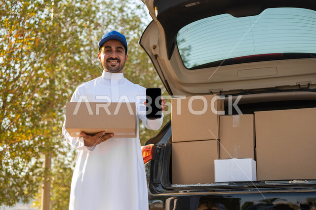 الشحن السريع للبضائع والمشتريات الأونلاين، عمليات النقل الموثوقة، مندوب توصيل يرتدي الثوب التقليدي وقبعة زرقاء يحمل بيده صندوق كرتون ويقوم بعرض شاشة سوداء فارغة عبر الجوال، التوصيل والشراء عن طريق تطبيقات الهاتف المحمول