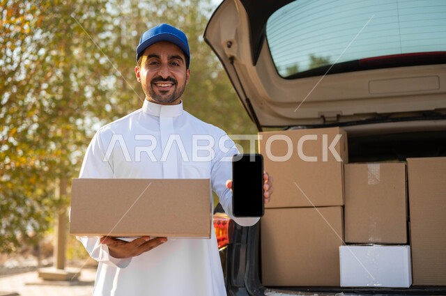 التوصيل والشراء عن طريق تطبيقات الهاتف المحمول، عمليات النقل الموثوقة، مندوب توصيل يرتدي الثوب التقليدي وقبعة زرقاء يحمل بيده صندوق كرتون ويقوم بعرض شاشة سوداء فارغة عبر الجوال، الشحن السريع للبضائع والمشتريات الأونلاين