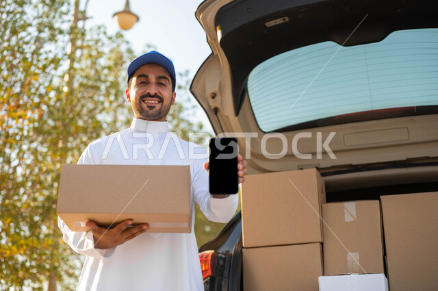 الشراء والتوصيل عن طريق تطبيقات الهاتف المحمول، عمليات النقل الموثوقة، مندوب توصيل يرتدي الثوب التقليدي وقبعة زرقاء يحمل بيده صندوق كرتون ويقوم بعرض شاشة سوداء فارغة عبر الجوال، الشحن السريع للبضائع والمشتريات الأونلاين