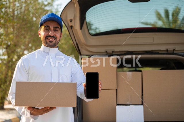 عمليات النقل الموثوقة، مندوب توصيل يرتدي الثوب التقليدي وقبعة زرقاء يحمل بيده صندوق كرتون ويقوم بعرض شاشة سوداء فارغة عبر الجوال، الشراء والتوصيل عن طريق تطبيقات الهاتف المحمول، الشحن السريع للبضائع والمشتريات الأونلاين