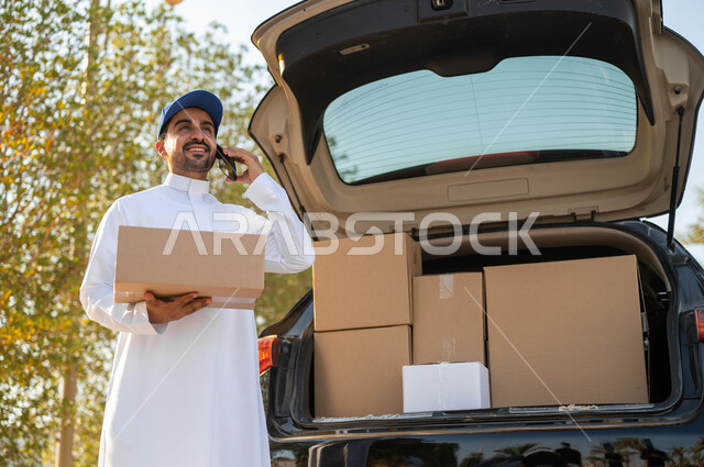عملية النقل السريع للطلبات والبضائع، التواصل الفعال مع العملاء، مندوب توصيل عربي خليجي سعودي مبتسم يرتدي الثوب التقليدي وقبعة زرقاء يقوم بإجراء مكالمة هاتفية، خدمات التوصيل الموثوقة