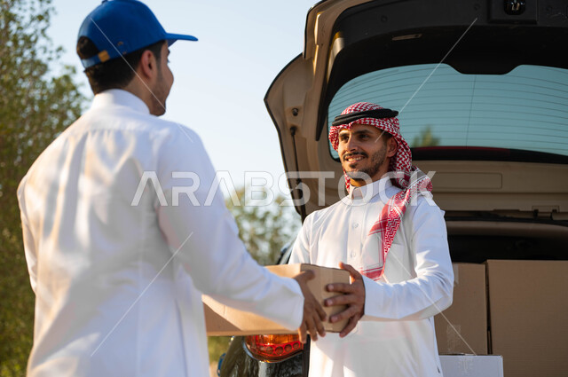 The process of transferring orders to customers, receiving the order from the goods vehicle, a Saudi Gulf Arab man wearing the traditional dress and shemagh taking the shopping box, receiving online orders, safe smart shopping, fast and reliable shipping service, ordering purchases online.