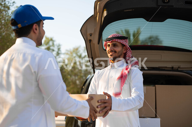 خدمة الشحن السريعة والموثوقة، استلام الطلبية من مندوب توصيل مبتسم يرتدي قبعة زرقاء، طلب المشتريات أونلاين، رجل عربي خليجي سعودي يرتدي الثوب التقليدي والشماغ يقوم بأخذ صندوق كرتون، عملية نقل الطلبات للزبائن
