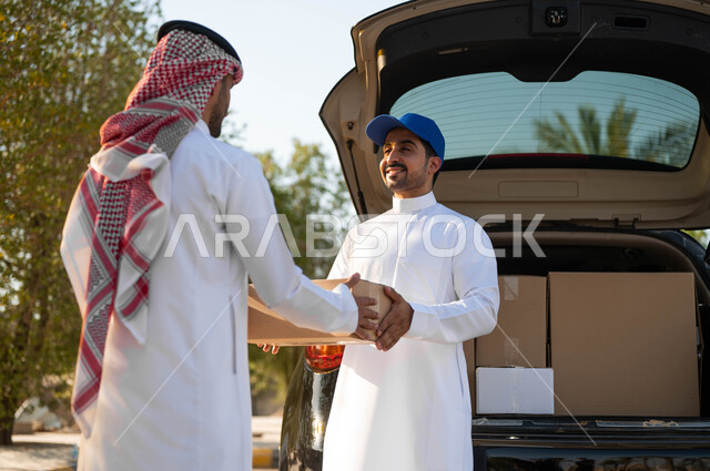 استلام الطلبية من مندوب توصيل مبتسم يرتدي قبعة زرقاء، طلب المشتريات أونلاين، رجل عربي خليجي سعودي يرتدي الثوب التقليدي والشماغ يقوم بأخذ صندوق كرتون، خدمة الشحن السريعة والموثوقة، عملية نقل الطلبات للزبائن