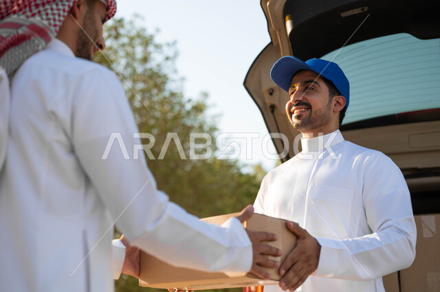 طلب المشتريات أونلاين، استلام الطلبية من مندوب توصيل مبتسم يرتدي قبعة زرقاء، صورة مقربة لرجل عربي خليجي سعودي يرتدي الثوب التقليدي والشماغ يقوم بأخذ صندوق كرتون، خدمة الشحن السريعة والموثوقة، عملية نقل الطلبات للزبائن