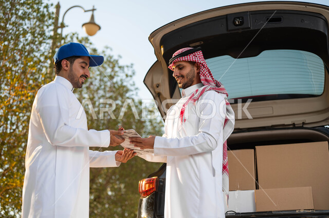ايماءات الرضا والسعادة لوصول الطرد بسرعة وسهولة، كراتين الشحن والتوصيل الأونلاين، صورة مقربة لشاب عربي سعودي خليجي يرتدي الثوب التقليدي والشماغ ويقوم بالتوقيع على استلام المشتريات