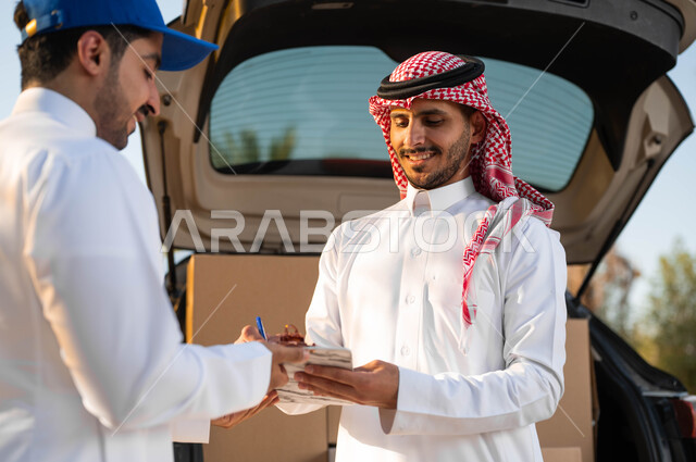 كراتين الشحن والتوصيل الأونلاين، ايماءات الرضا والسعادة لوصول الطرد بسرعة وسهولة، صورة مقربة لشاب عربي سعودي خليجي يرتدي الثوب التقليدي والشماغ يقوم بالتوقيع على استلام المشتريات