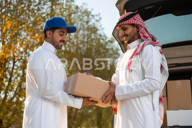 توصيل الطلبات الى باب المنزل من خلال خدمة الديلفري، صورة مقربة لشاب عربي سعودي خليجي يرتدي الزي التقليدي يضع على رأسه قبعة شمسية زرقاء يقوم بتسليم الطرد للزبون، ايماءات الرضا والسعادة، التوقيع على استلام الطلبية للتأكيد على سلامتها