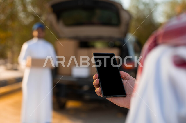 Online shipping and delivery cartons arrive, a Saudi Arabian Gulf man holds a mobile phone while receiving a delivery order, shipping orders to the door of the house quickly and easily, shopping and ordering products online