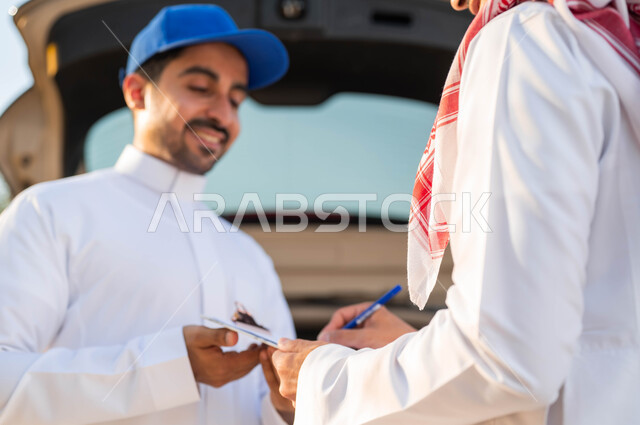 التوقيع على أوراق و مستندات للتأكيد على استلام و تسليم الطرد، صورة مقربة لشابان عربيين خليجيين سعوديين يرتديان الزي التقليدي يقفان أمام سيارة التوصيل، الحصول على الطلبات بسرعة وسهولة، ايماءات الرضا والسعادة