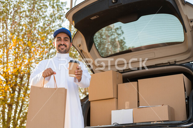 توصيل الطلبات للزبائن، الوقوف بجانب سياره شحن البضائع، رجل عربي سعودي خليجي مبتسم يرتدي الزي التقليدي يضع على راسه قبعه شمسيه زرقاء ويحمل بيده كوب قهوة وكيس تسوق ورقي، خدمات التوصيل السريع والآمن للمشروبات الساخنه