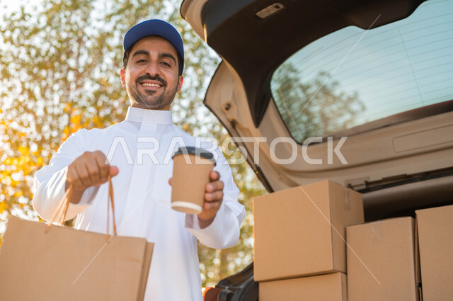 الوقوف بجانب سياره شحن البضائع ,خدمات التوصيل السريع والآمن للمشروبات الساخنة، صورة مقربة لمندوب عربي سعودي خليجي يرتدي الزي التقليدي و القبعة الزرقاء يحمل بيده كوب قهوة وكيس تسوق ورقي، النظر إلى الكاميرا بإيماءات الفرح