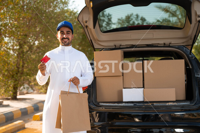 عملية شحن الطلبات السريعة الى المنزل، الوقوف بجانب سياره البضائع، رجل عربي سعودي خليجي يرتدي الزي التقليدي و القبعة الزرقاء ويحمل بيده كيس تسوق ورقي و بطاقة الدفع الالكتروني، خدمات التوصيل الآمن, النظر إلى الكاميرا بإيماءات الفرح.