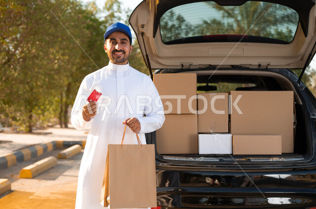 خدمات الشحن والتوصيل ذات الجودة العالية، الوقوف بجانب سياره شحن البضائع، رجل عربي سعودي خليجي يرتدي الزي التقليدي و القبعة الزرقاء ويحمل بيده كيس تسوق ورقي و بطاقة الدفع الالكتروني، خدمات التوصيل السريع و الآمن, النظر إلى الكاميرا بإيماءات السرور.