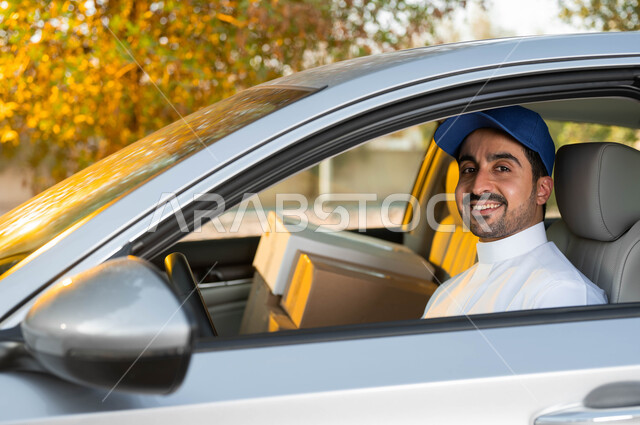 High-quality shipping services in the Kingdom of Saudi Arabia, a close-up picture of a smiling Saudi Arabian Gulf delivery representative wearing a traditional dress and a blue hat, looking at the camera with an expression of pleasure and happiness, sitting inside a goods shipping vehicle