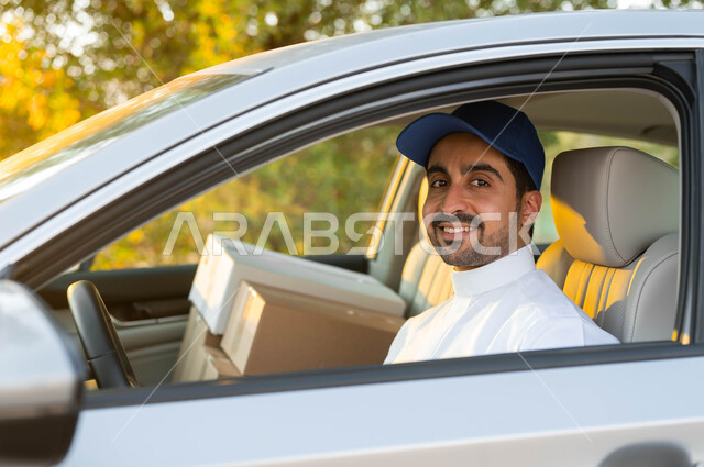 خدمات الشحن عالية الجودة في المملكة السعودية, صورة مقربة لمندوب توصيل عربي خليجي سعودي مبتسم يرتدي الزي التقليدي و القبعة الزرقاء, الجلوس داخل سيارة شحن البضائع, النظر إلى الكاميرا بإيماءات الرضا و السرور.