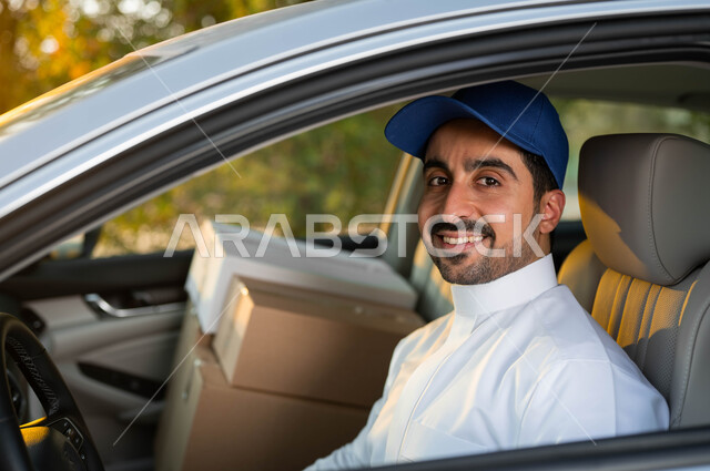 الجلوس داخل سيارة شحن البضائع, صورة مقربة لمندوب عربي خليجي سعودي مبتسم يرتدي الزي التقليدي و القبعة الزرقاء ينظر إلى الكاميرا بإيماءات الفرح و السرور, خدمات التوصيل السريعة في السعودية, عملية نقل الطلبات للزبائن, استخدام التقنيات الحديثة المتطورة